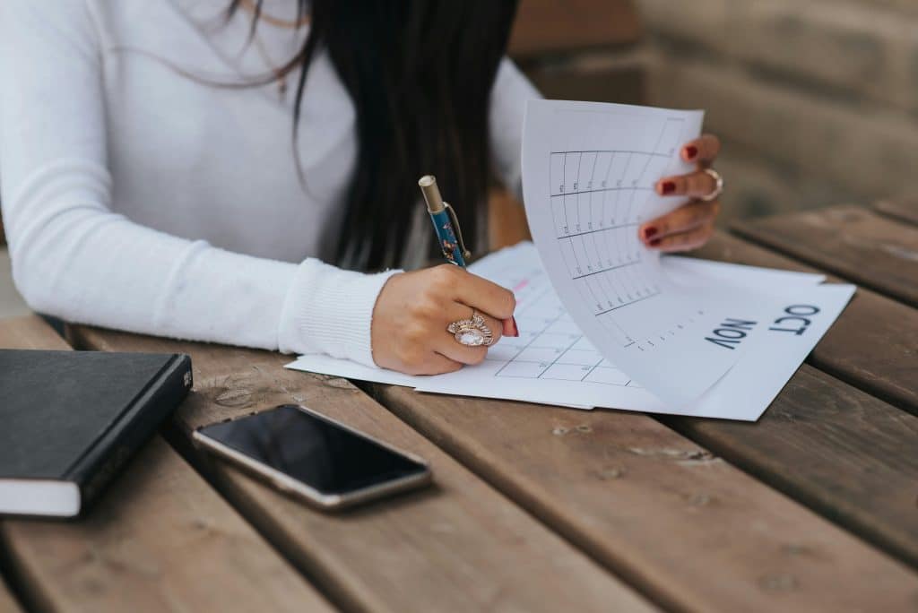 Une femme assise devant une table avec son agenda et son téléphone posé à côté, en train de remplir son agenda à un jour précis.